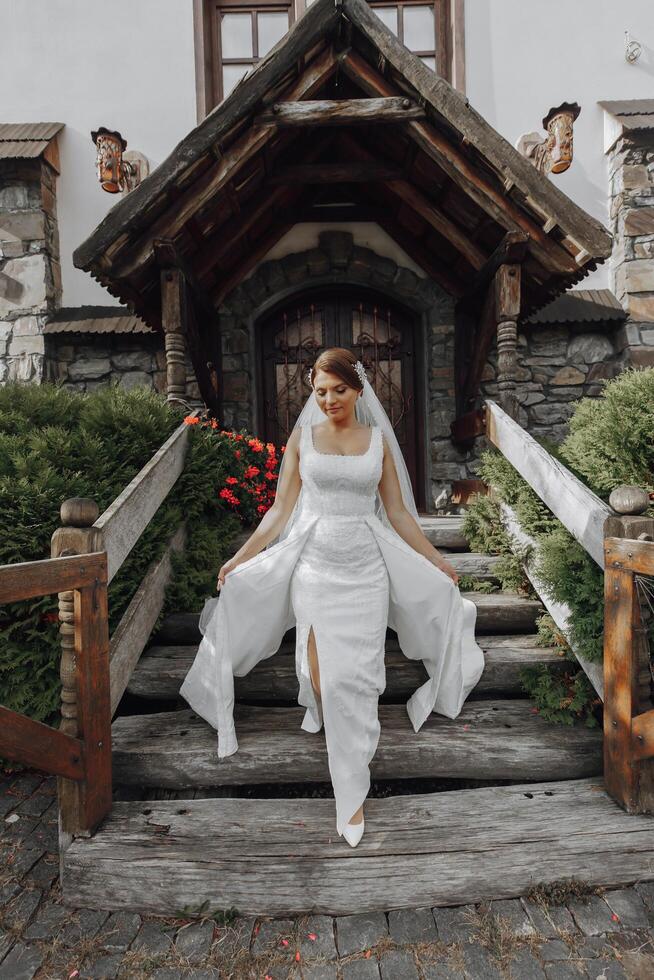 romantisch in voller Länge Porträt von ein braunhaarig Frau im ein Weiß Kleid mit ein Strauß Stehen in der Nähe von das hölzern Tor von das Kirche Eingang foto
