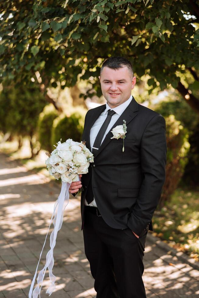 Porträt von ein gut aussehend jung Bräutigam auf ein Herbst Tag draußen im ein elegant passen und halten ein Hochzeit Strauß von Blumen. draussen Foto. gut aussehend Mann im ein Geschäft Anzug. foto