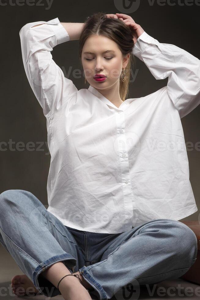 schön Mädchen im ein Weiß Hemd und Jeans posieren auf ein grau Hintergrund. foto