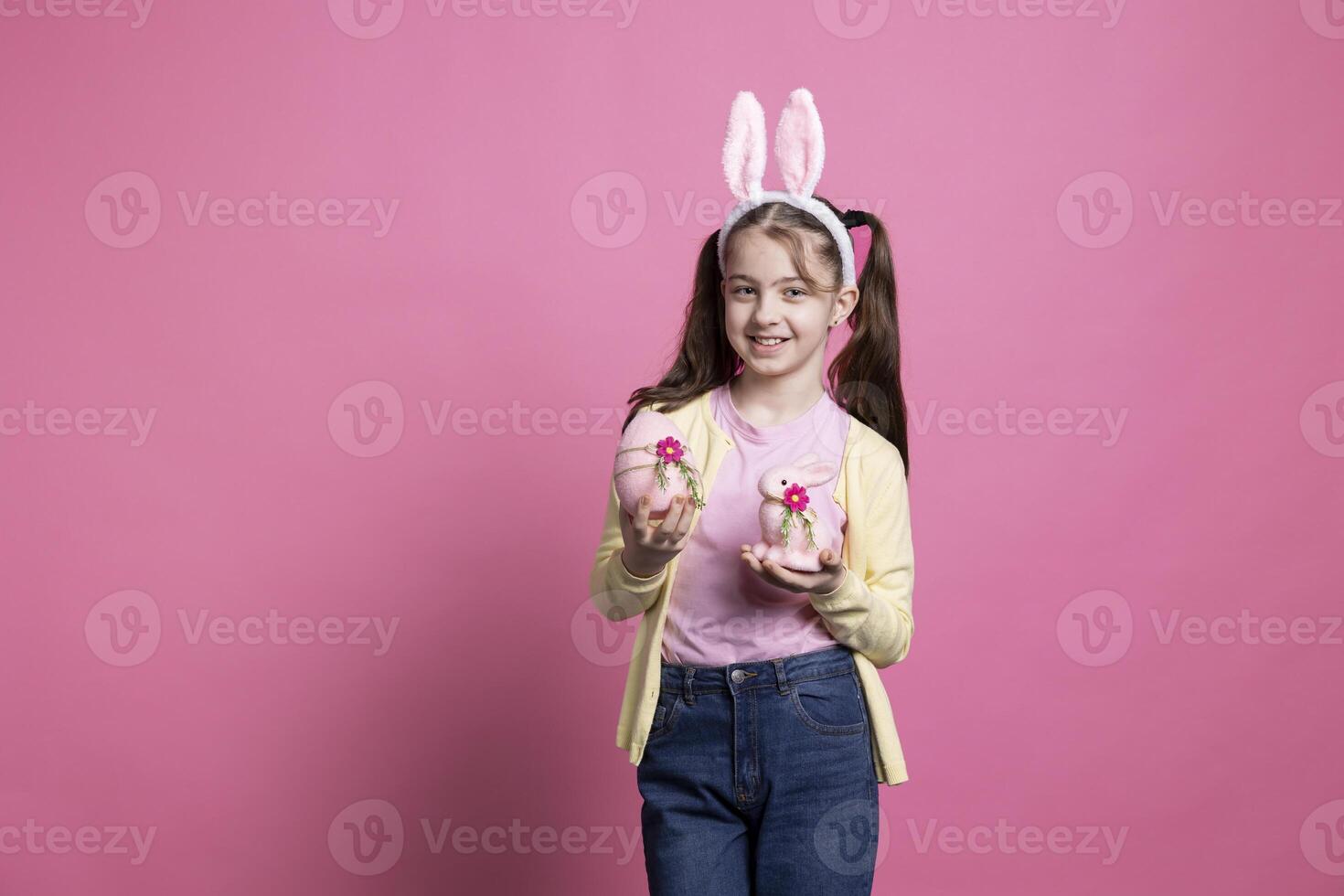 wenig Mädchen mit flauschige Hase Ohren hält Rosa Ostern Vereinbarungen im Vorderseite von Kamera, zeigen ihr dekoriert Ei und ausgestopft Hase Spielzeug. jung Kind lächelnd im Studio und feiern April Fall. foto