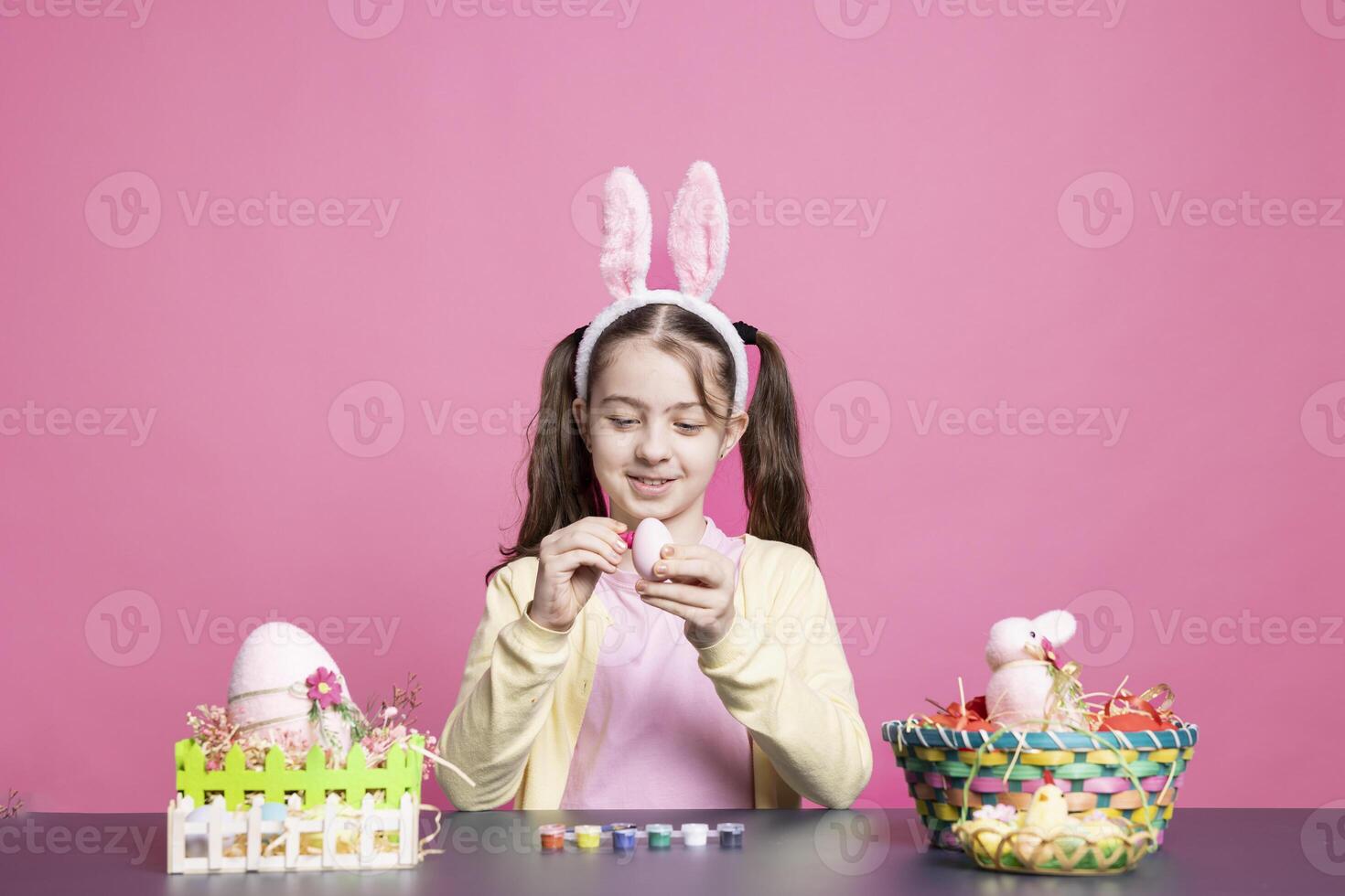 heiter Schülerin mit Hase Ohren Gemälde Eier beim ein Tisch, Erstellen handgemacht festlich Dekorationen zum Ostern Feier Tradition. jung lächelnd Mädchen genießt dekorieren Ornamente mit Krawatte Farbstoff. foto