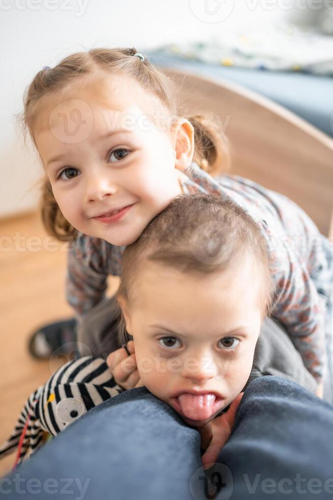 klein Junge mit Nieder Syndrom Theaterstücke mit seine jünger Schwester im Zuhause Schlafzimmer. hoch Qualität Foto