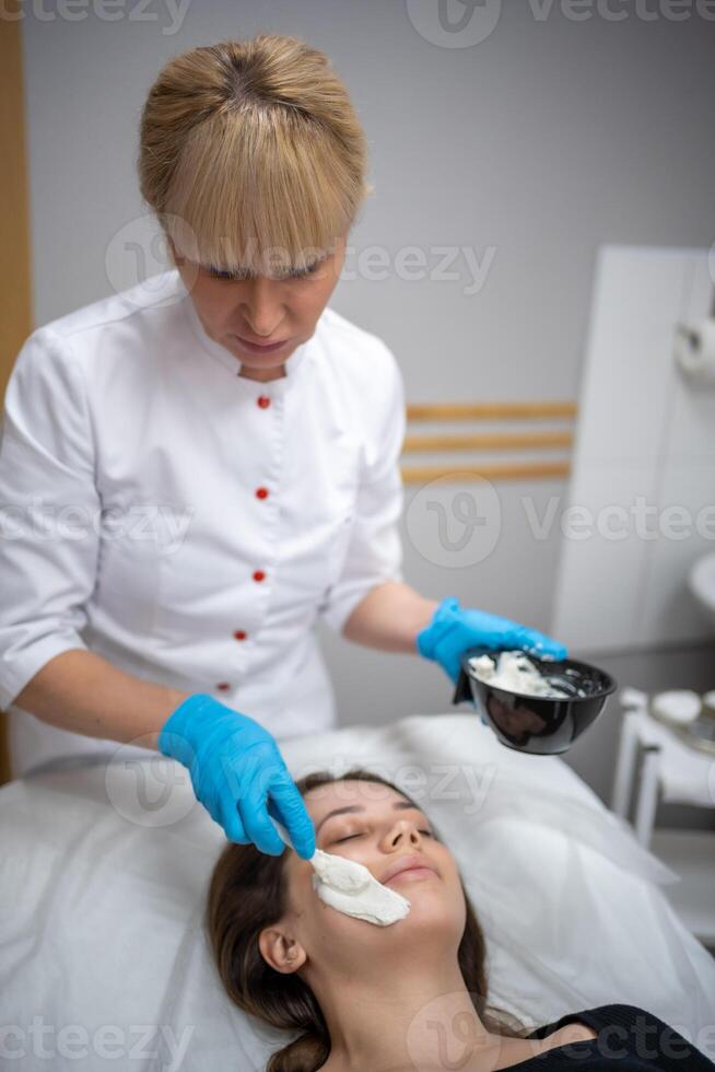 Arzt Kosmetikerin oder Dermatologe Herstellung Gesicht Maske im Kosmetologie Salon. Fachmann Kosmetikerin bewirbt sich Gesicht Maske auf kaukasisch Frau Gesicht Lügen auf Bett im Bademantel. foto