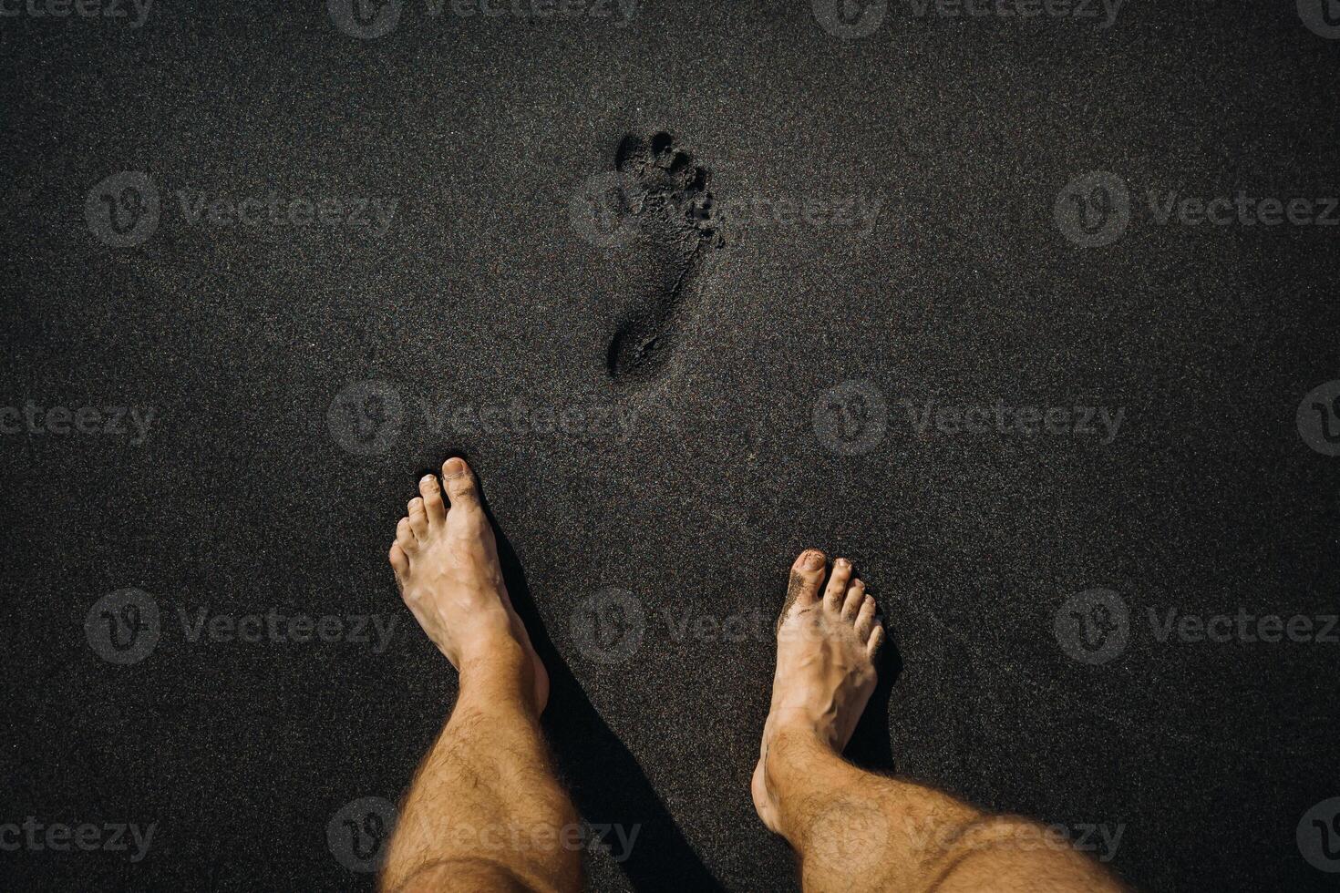 schließen oben von männlich Fußabdrücke und Füße Gehen auf das vulkanisch schwarz Sand auf das Strand foto