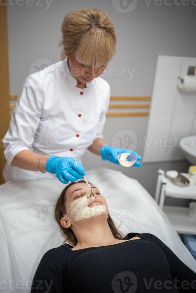 Arzt Kosmetikerin oder Dermatologe Herstellung Gesicht Maske im Kosmetologie Salon. Fachmann Kosmetikerin bewirbt sich Gesicht Maske auf kaukasisch Frau Gesicht Lügen auf Bett im Bademantel. foto