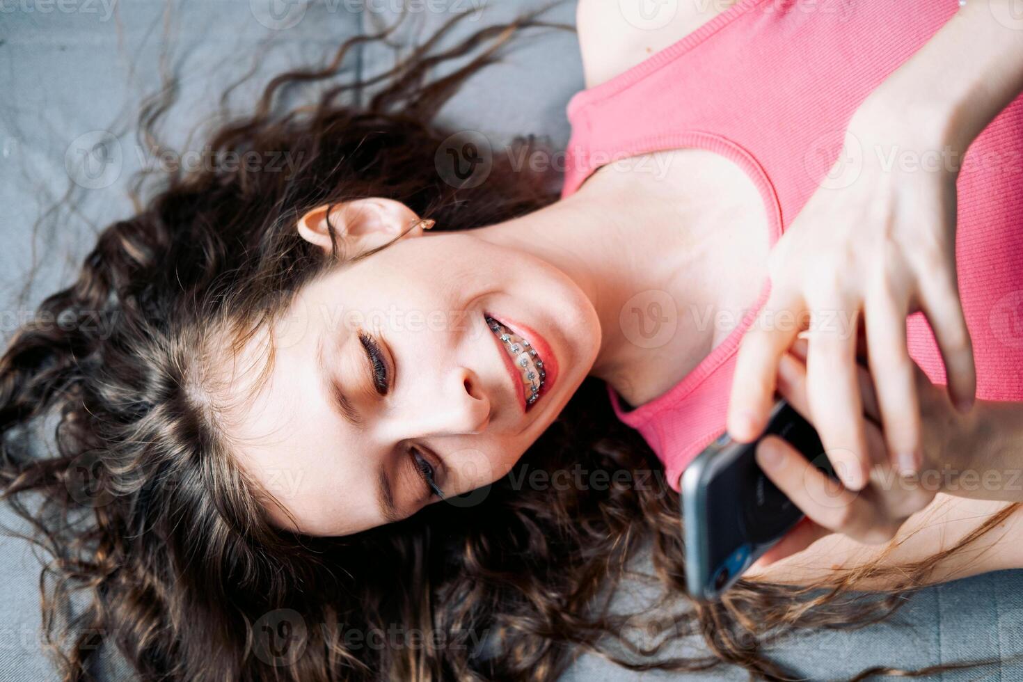 jung Frau im Hosenträger mit lange lockig Haar Lügen auf das Sofa und Schriftrollen Sozial Netzwerke auf ihr Telefon im ein Rosa Spitze, oben Aussicht foto