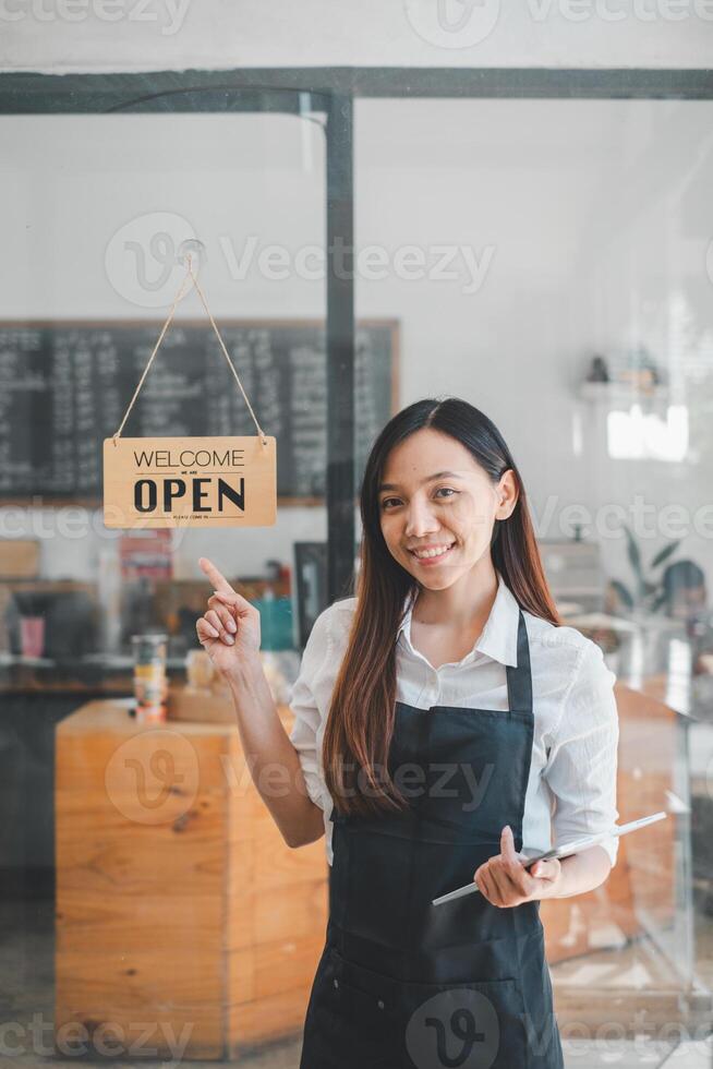 Porträt von glücklich Kellnerin Stehen beim Restaurant Eingang mit öffnen Zeichen, Porträt von jung Geschäft Frau besuchen Neu Kunden im ihr Kaffee Geschäft. foto
