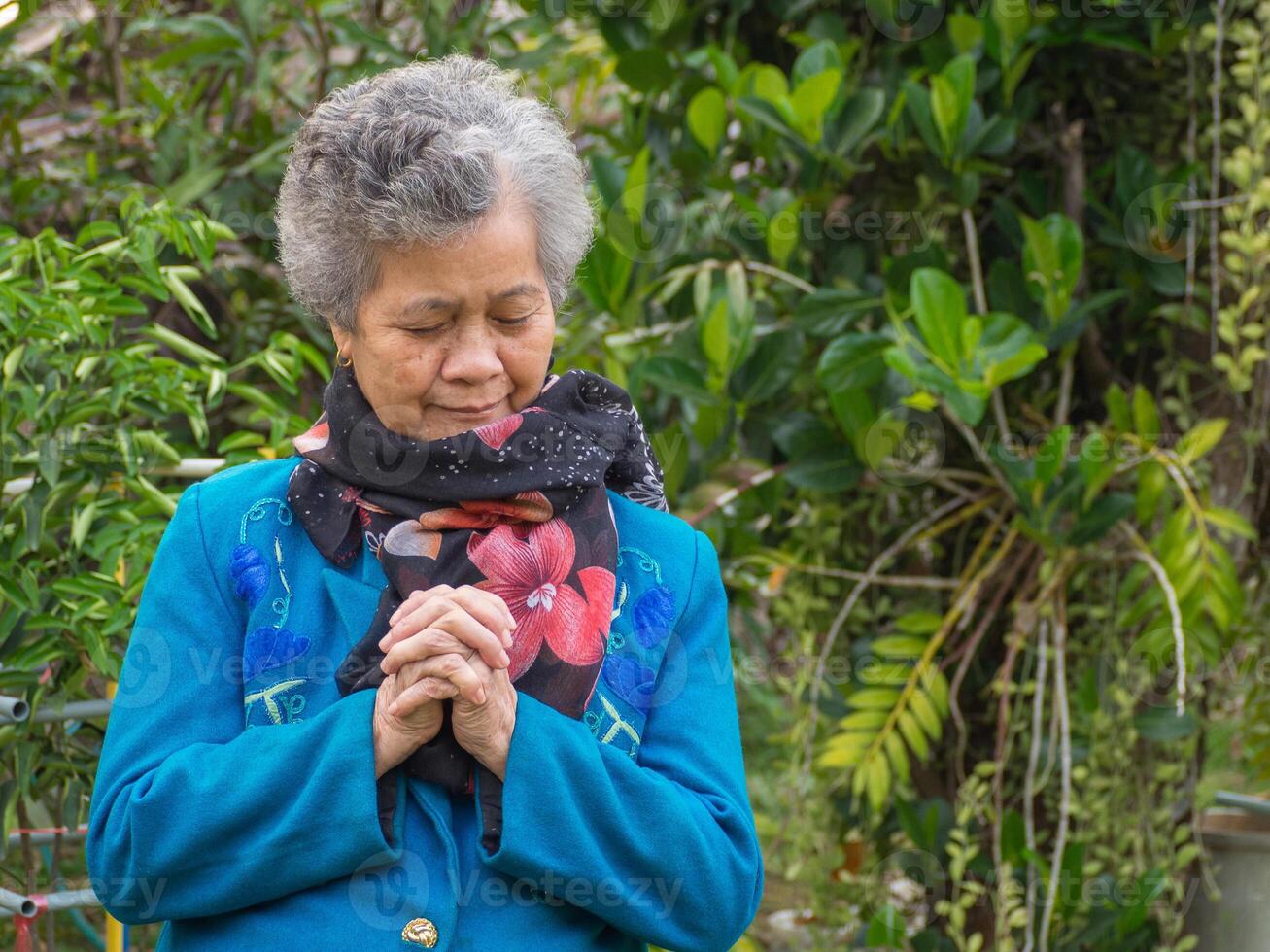 ein Alten asiatisch Frau beten und geschlossen Augen während Stehen im ein Garten. Raum zum Text. Konzept von alt Menschen und Gesundheitswesen foto