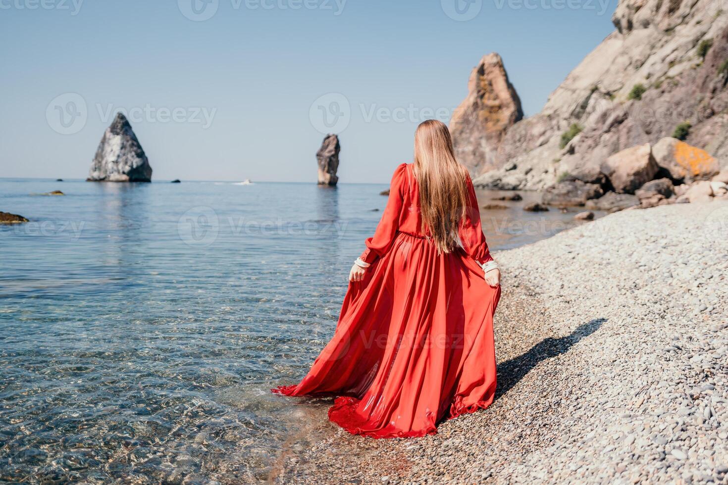 Frau Reise Meer. glücklich Tourist im rot Kleid genießen nehmen Bild draußen zum Erinnerungen. Frau Reisender posieren auf das Felsen beim Meer Bucht umgeben durch vulkanisch Berge, Teilen Reise Abenteuer Reise foto