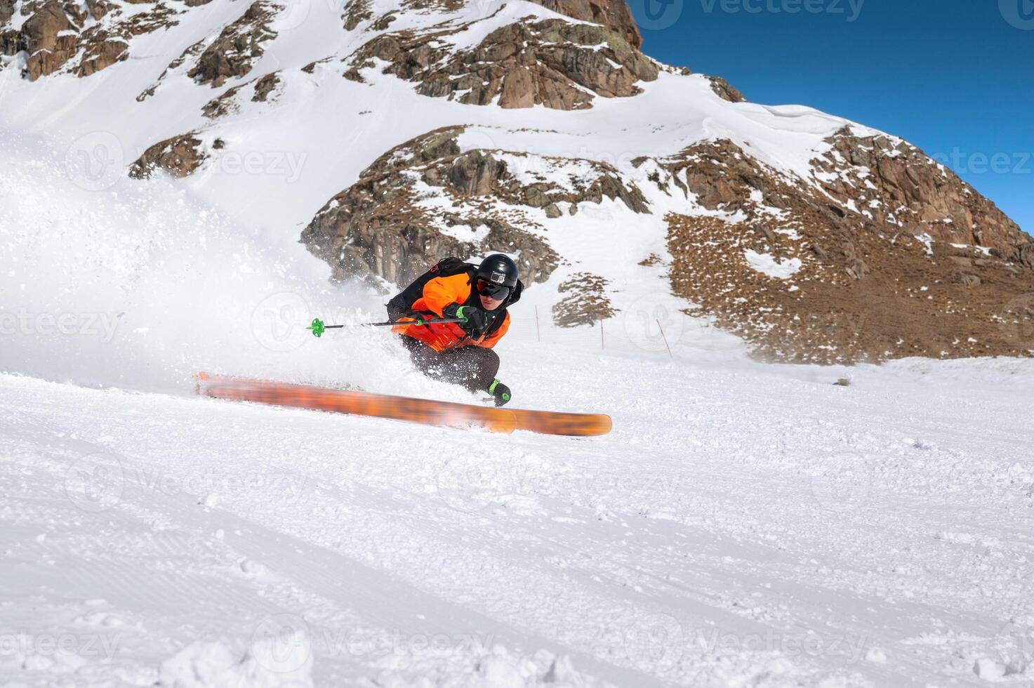 ein Skifahrer im ein Orange Jacke und ein schwarz Helm mit ein Maske Fahrten Nieder ein schneebedeckt Steigung beim hoch Geschwindigkeit. Pulver von unter Ski und ein aussehen beim das Kamera foto