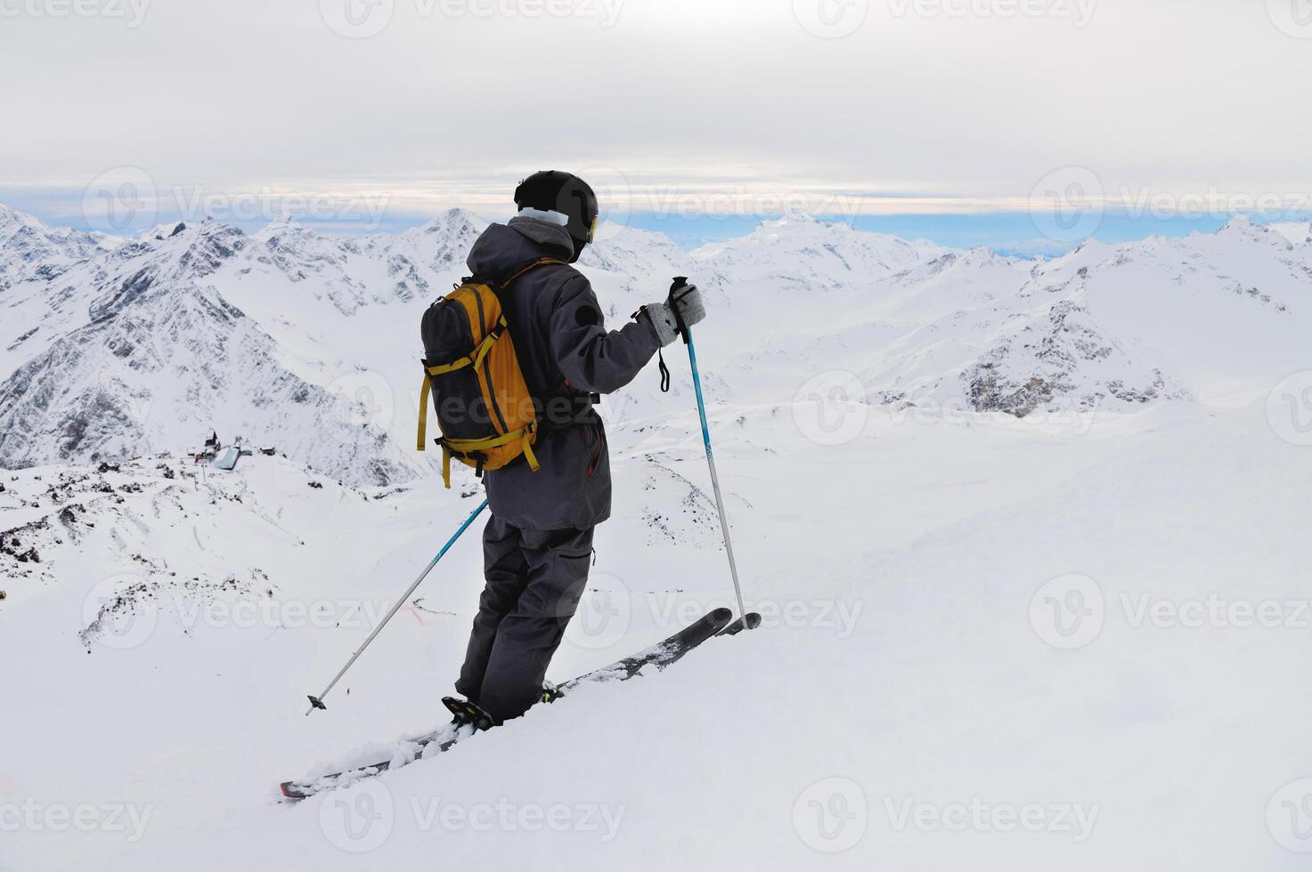 Seite Aussicht von ein sportlich Skifahrer im ein Helm und Brille mit ein Rucksack, Stehen auf Ski, halten Ski Stangen im Weiß Schnee, gegen das Himmel, genießen ein schön Aussicht von das Berge foto