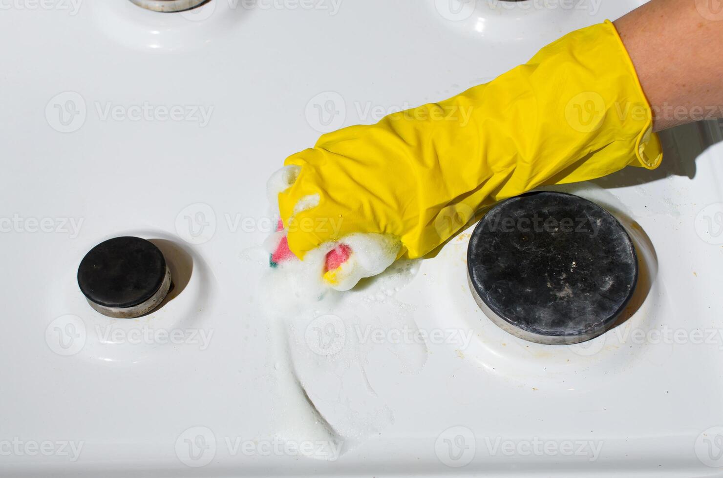 weiblich Hand im Handschuh Reinigung das Oberfläche von Gas Herd mit Waschen Schwamm und chemisch Schaum. oben Sicht. Zuhause Reinigung Konzept, Küche Hygiene foto