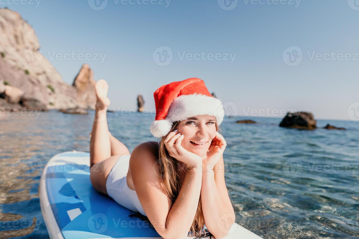 Frau Meer sup. schließen oben Porträt von glücklich jung kaukasisch Frau mit lange Haar im Santa Hut suchen beim Kamera und lächelnd. süß Frau Porträt im ein Weiß Bikini posieren auf sup Tafel im das Meer foto