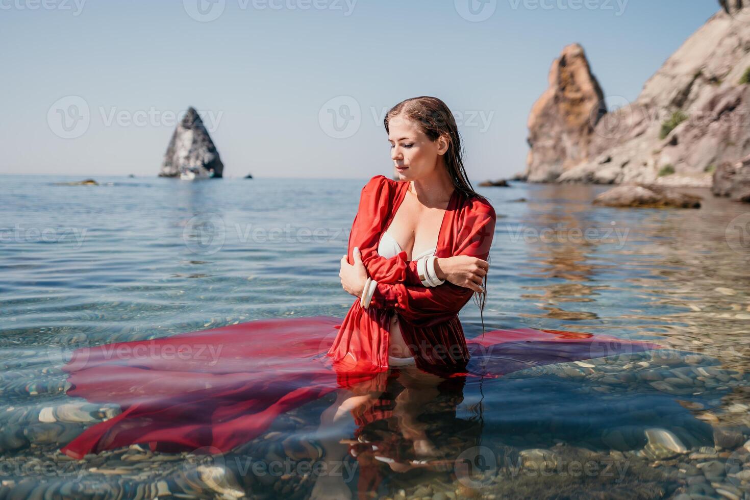 Frau Reise Meer. glücklich Tourist im rot Kleid genießen nehmen Bild draußen zum Erinnerungen. Frau Reisender posieren im Meer Strand, umgeben durch vulkanisch Berge, Teilen Reise Abenteuer Reise foto