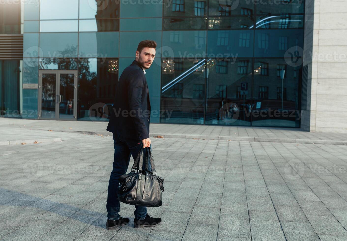 gut aussehend jung Geschäftsmann mit ein Bart und im ein Geschäft passen Stehen auf das Straße gegen das Hintergrund von das Büro Gebäude Nächster zu ein komfortabel stilvoll Leder Tasche. foto