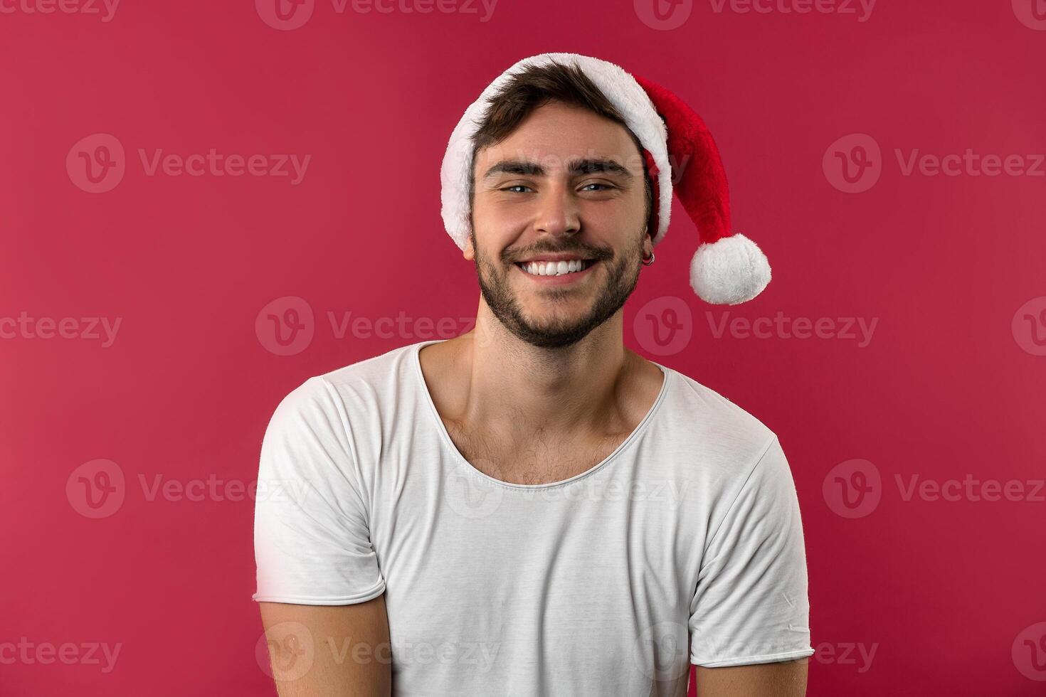 jung gut aussehend kaukasisch Kerl im ein Weiß T-Shirt und Santa Hüte steht auf rot Hintergrund im Studio und und Zähne lächelnd foto