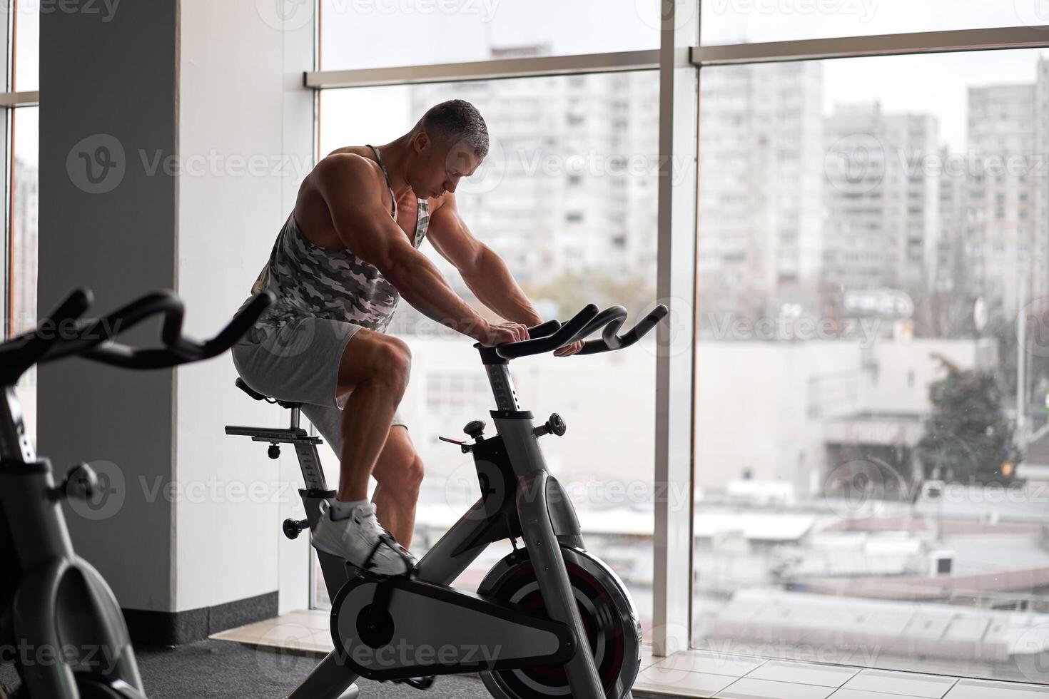 muskulös sportlich Bodybuilder Fitness Modell- Sitzung stationär Fahrrad im Fitnessstudio in der Nähe von groß Fenster foto