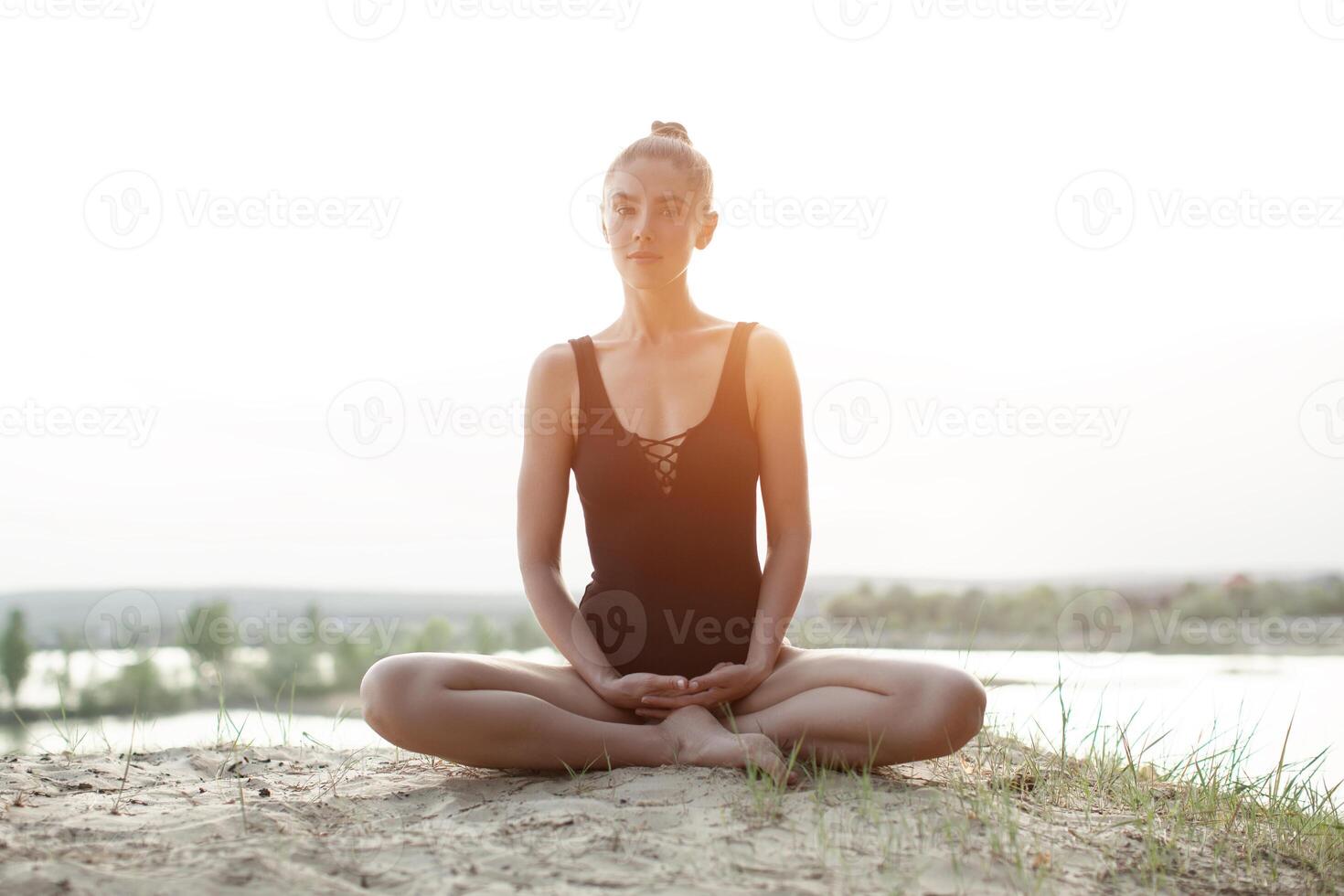 ein schön kaukasisch Mädchen im ein schwarz Badeanzug ist Sitzung im das Lotus Position auf Natur gegen das Hintergrund von das hell Sonne. foto