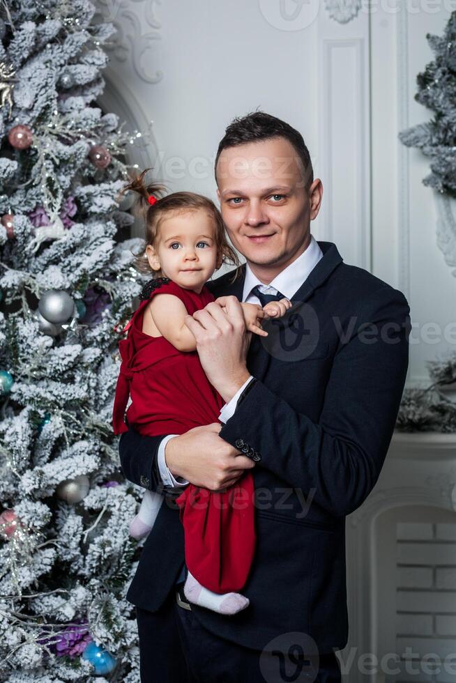 Papa im ein Geschäft passen hält seine Tochter gekleidet im ein elegant rot Kleid auf das Hintergrund von das Weihnachten Baum foto