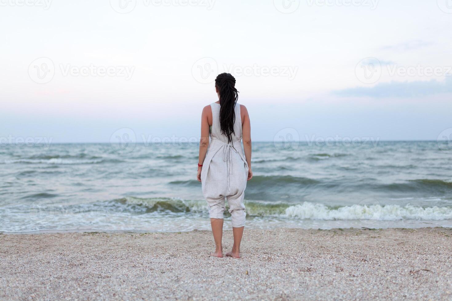zurück Aussicht von schön Frau im Weiß Kleid mit Dreadlock auf das Kopf genießen das idyllisch Szene auf das Strand. foto
