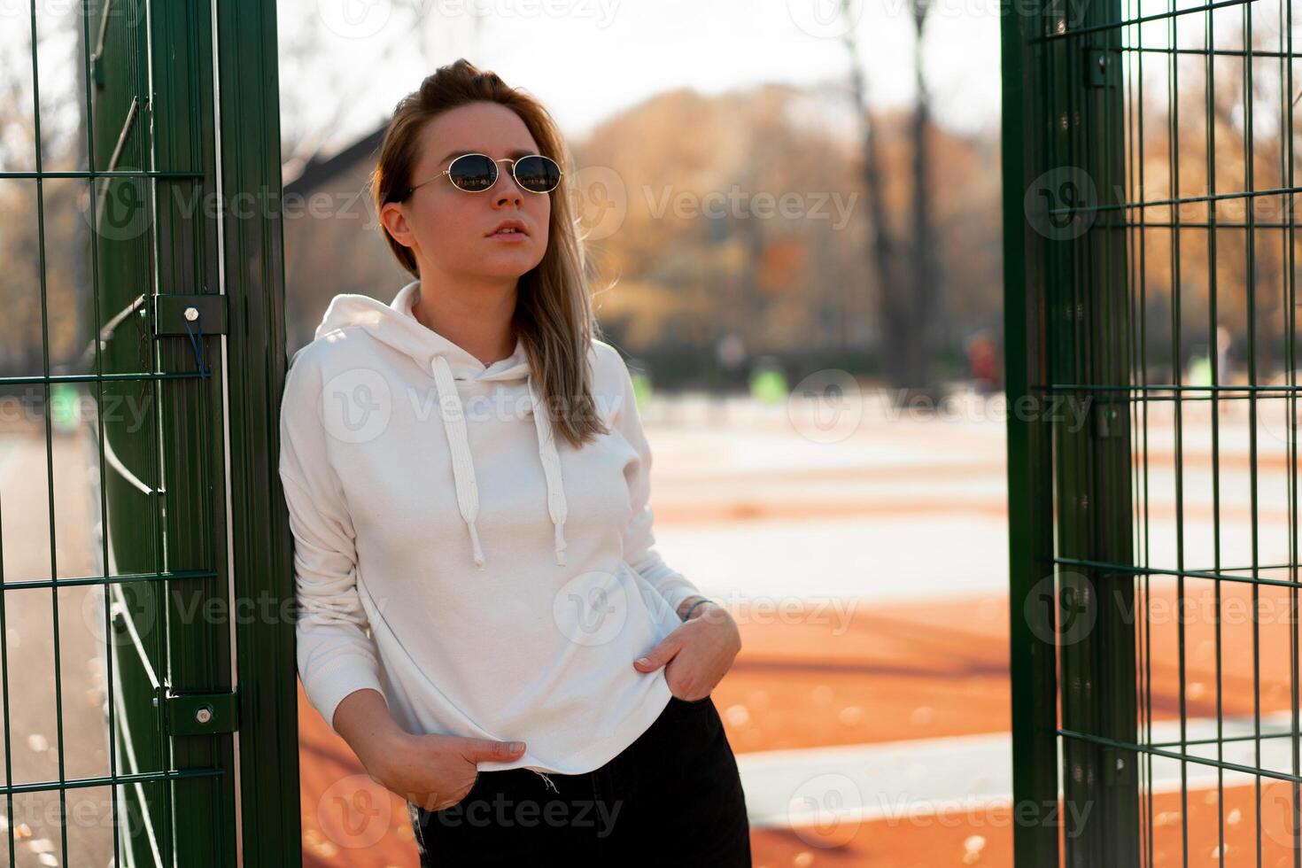 draussen schließen oben Porträt von jung schön Frau mit lange Haar im Sonnenbrille, gekleidet im ein Weiß Pullover, in der Nähe von das Sportplatz foto