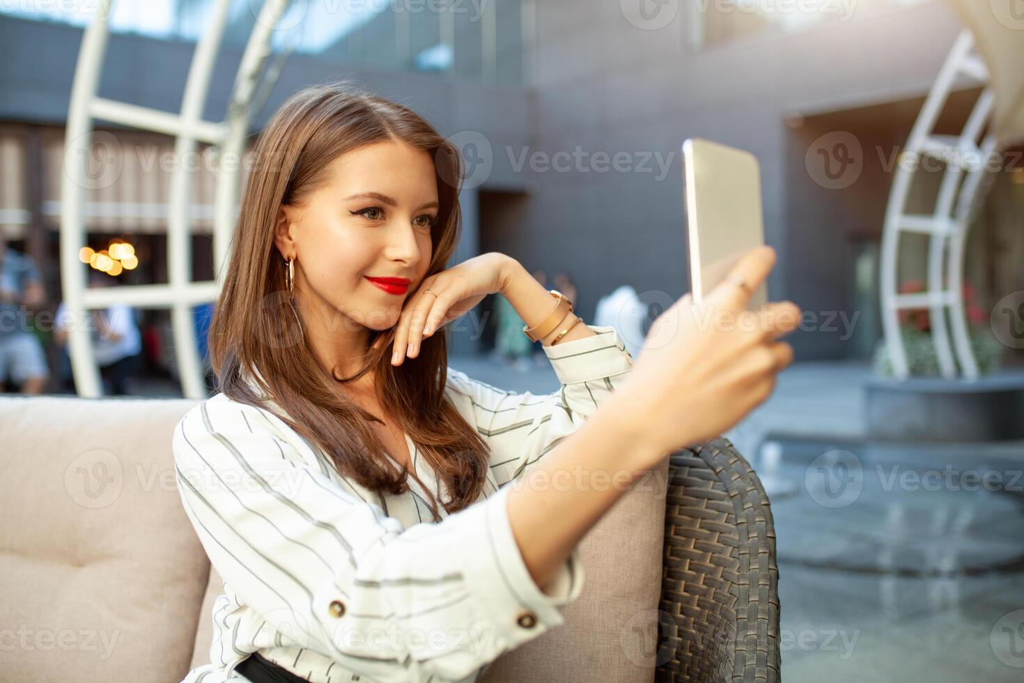 ein jung schön Mädchen gekleidet im ein gestreift modisch Kleid ist Sitzung im ein Sommer- draussen Cafe und ist warten zum ihr bestellen und ihr freunde oder ihr Freund. druckt ein Botschaft auf das Telefon foto