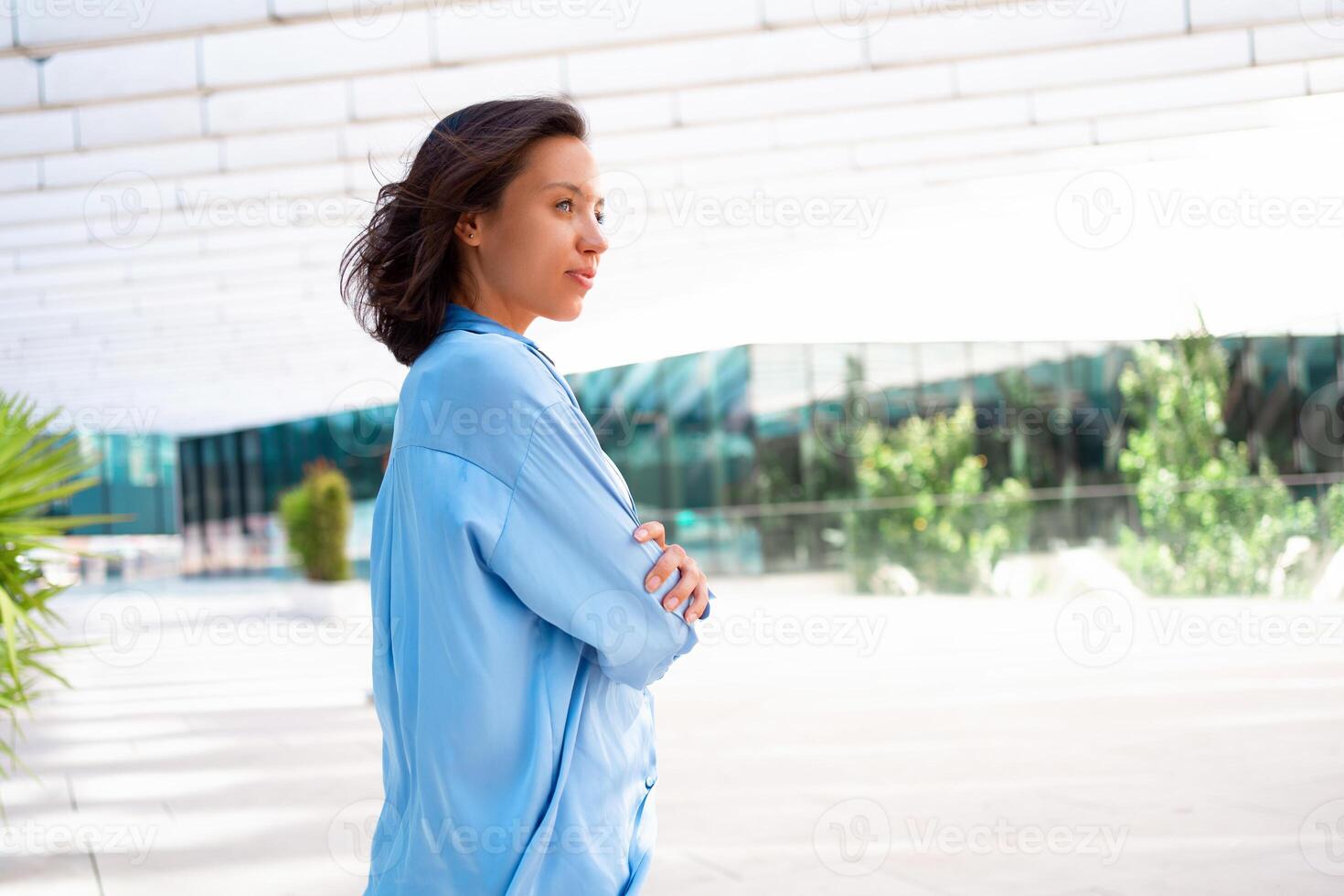 Vertrauen Geschäftsfrau Porträt mit gekreuzt Hände. ziemlich Geschäft Frau 30 Jahre alt Stehen in der Nähe von Büro Gebäude gekleidet Blau Shirt. foto