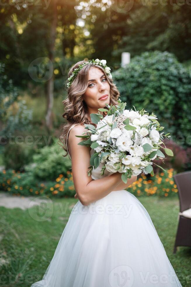 ein süß lockig Frau im ein Weiß Hochzeit Kleid mit ein Hochzeit Strauß und Kranz im ihr Haar Stehen zurück zu das Kamera im Natur. Konzept entkam Braut. nach vorne zu ein glücklich hell Zukunft weglaufen foto