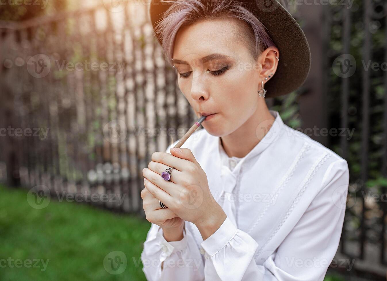jung stilvoll Hipster Frau tragen das Hut Stehen in der Nähe von das Zaun und Rauchen ein Zigarette im das Stadt foto