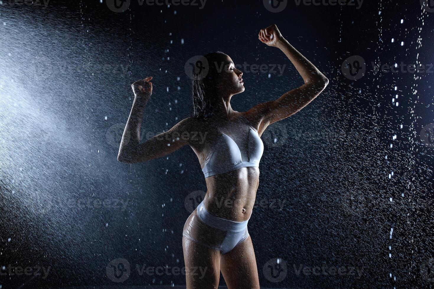 schön jung Mädchen im Sportbekleidung posiert im aqua Studio. Tropfen von Wasser Verbreitung Über ihr Fitness Körper. das perfekt Zahl auf das Hintergrund von Wasser spritzt foto