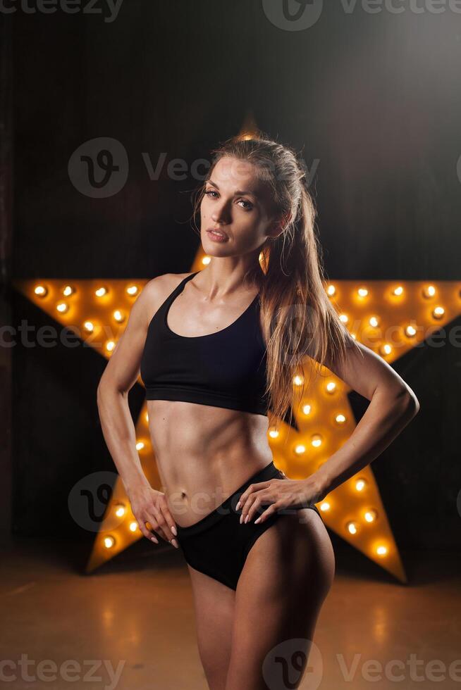 Bild von muskulös jung weiblich Athlet tragen schwarz Sport tragen Stehen in der Nähe von das Mauer auf dunkel Hintergrund. Frau Bodybuilder ruhen nach trainieren. foto