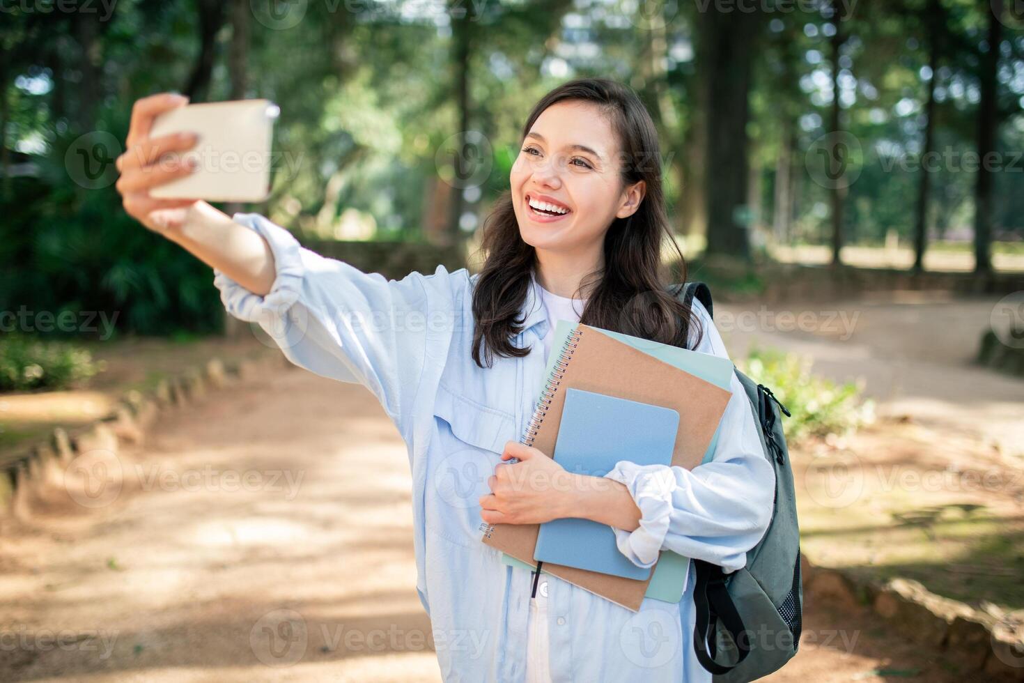 überschwänglich jung Frau nehmen ein Selfie mit ihr Smartphone während halten Lehrbücher foto