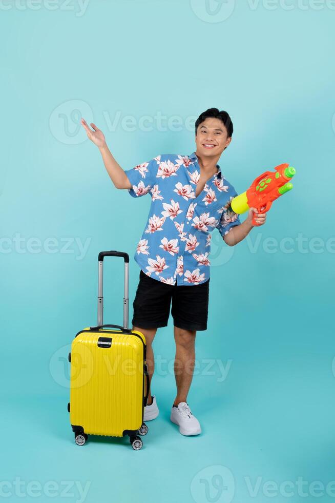 voll Körper Foto von gut aussehend asiatisch Tourist im Sommer- Kleider mit Wasser Gewehr und Gepäck während Songkran Festival auf Blau Hintergrund mit Kopieren Raum.