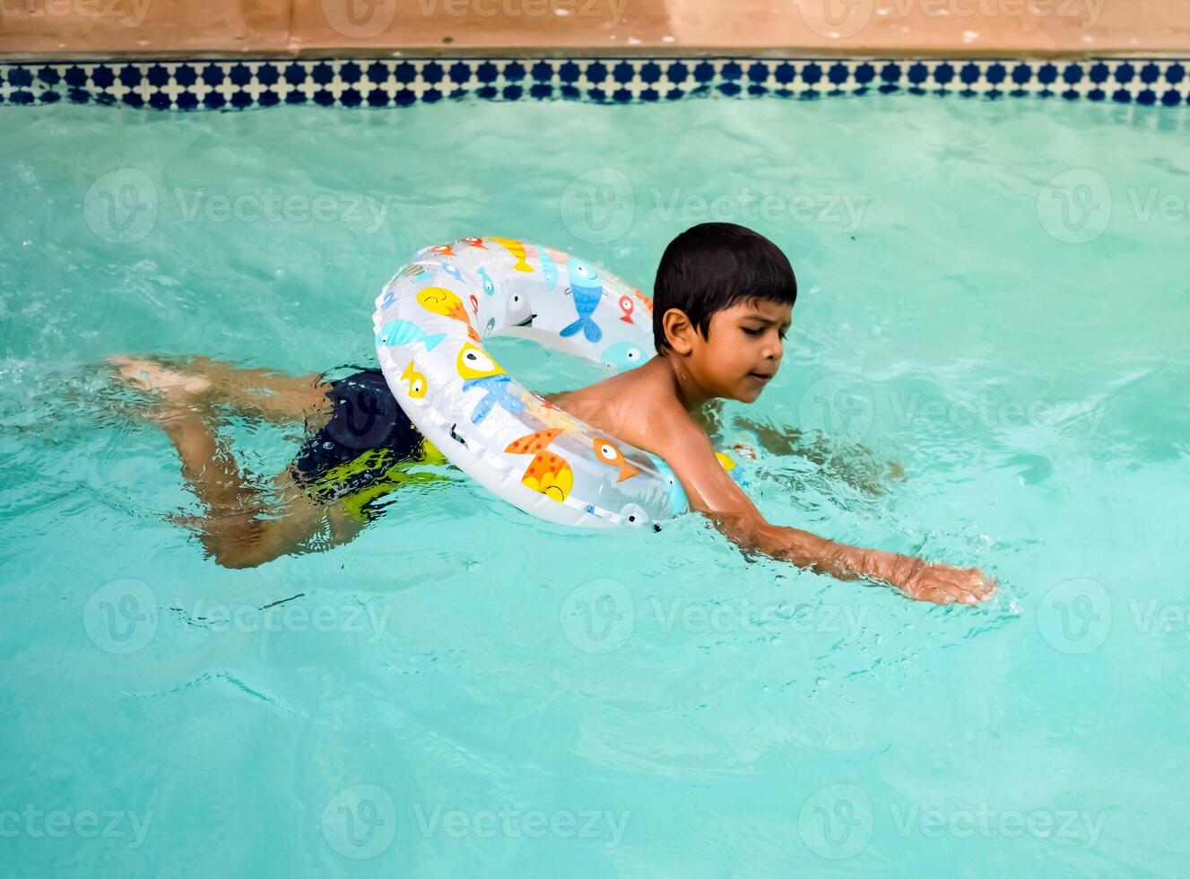 glücklich indisch Junge Schwimmen im ein Schwimmbad, Kind tragen Schwimmen Kostüm entlang mit Luft Tube während heiß Sommer- Urlaube, Kinder Junge im groß Schwimmen Schwimmbad. foto