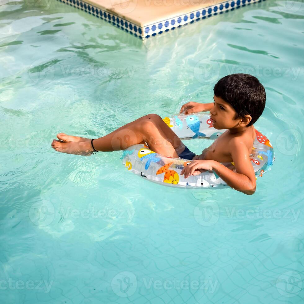 glücklich indisch Junge Schwimmen im ein Schwimmbad, Kind tragen Schwimmen Kostüm entlang mit Luft Tube während heiß Sommer- Urlaube, Kinder Junge im groß Schwimmen Schwimmbad. foto