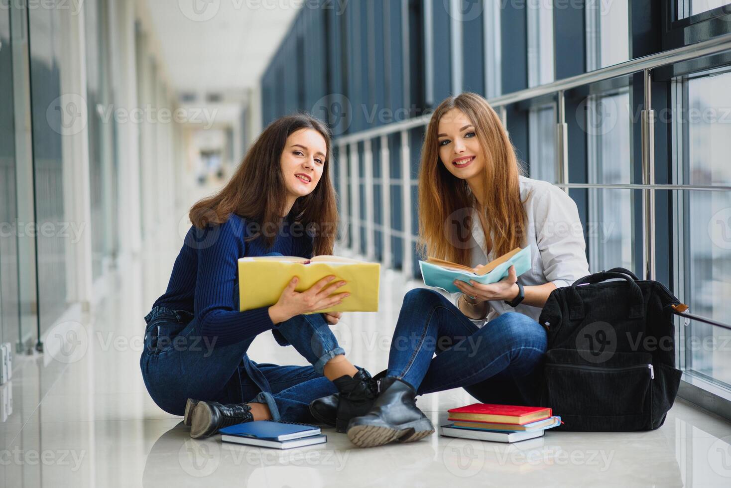 zwei ziemlich weiblich Studenten mit Bücher Sitzung auf das Fußboden im das Universität Flur foto