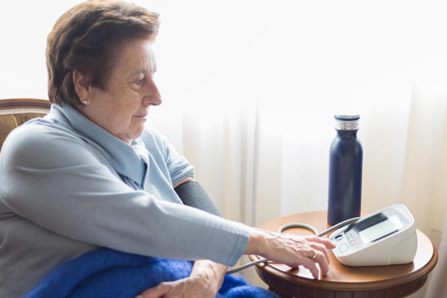 Kaukasische ältere Frau, die ihren Blutdruck zu Hause mit einem digitalen Monitor überprüft. kurzes Haar, blauer Pullover, Decke, kaltes Wasser foto