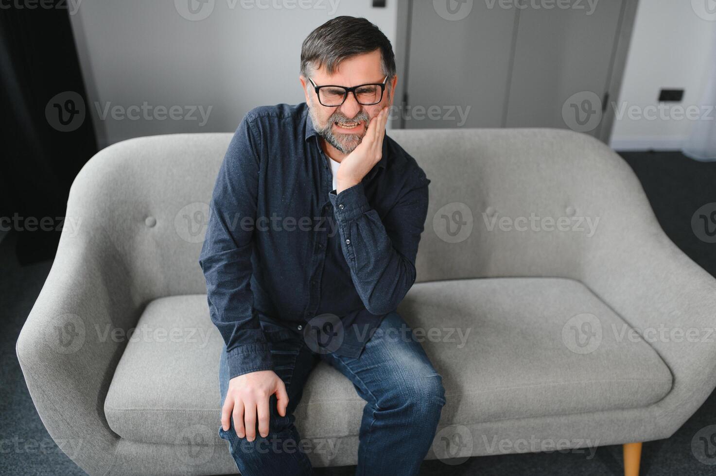 Alten Mann haben Zahnschmerzen berühren Wange Leiden von Schmerzen Sitzung auf Sofa beim heim. selektiv Fokus foto