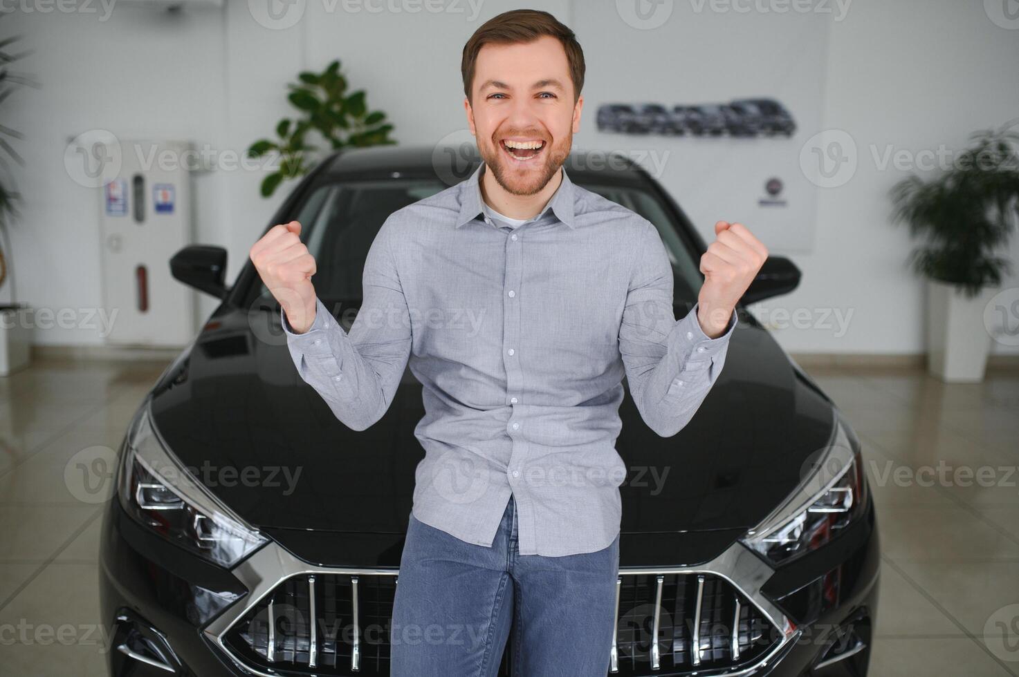 emotional gut aussehend Kerl erziehen Hand oben und lächelnd beim Kamera. Reich Mann Kauf Auto beim Luxus Händler Salon foto