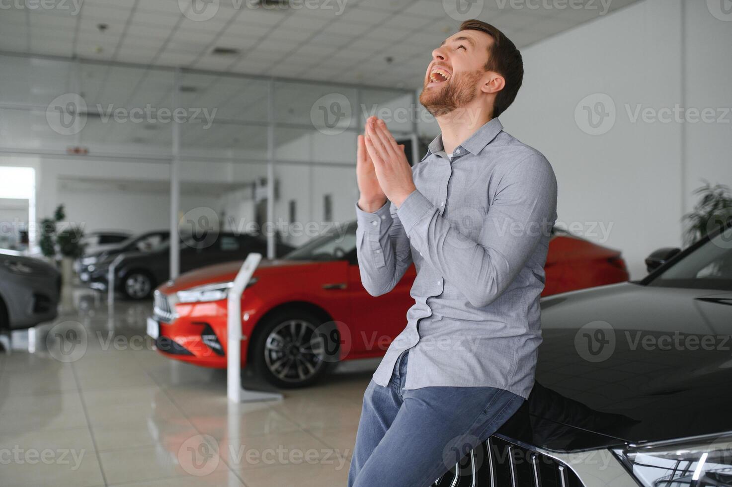 emotional gut aussehend Kerl erziehen Hand oben und lächelnd beim Kamera. Reich Mann Kauf Auto beim Luxus Händler Salon foto