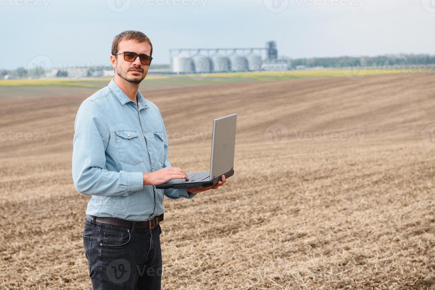 modern Farmer Überprüfung seine Feld Pflanze und Arbeiten auf Laptop Computer gegen Mais Trockner Silos im Konzept von industriell und Landwirtschaft foto