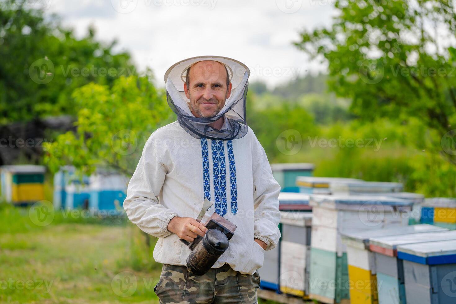 Imker im schützend Arbeitskleidung. Nesselsucht Hintergrund beim Bienenhaus. funktioniert auf das Bienenstände im das Frühling. foto