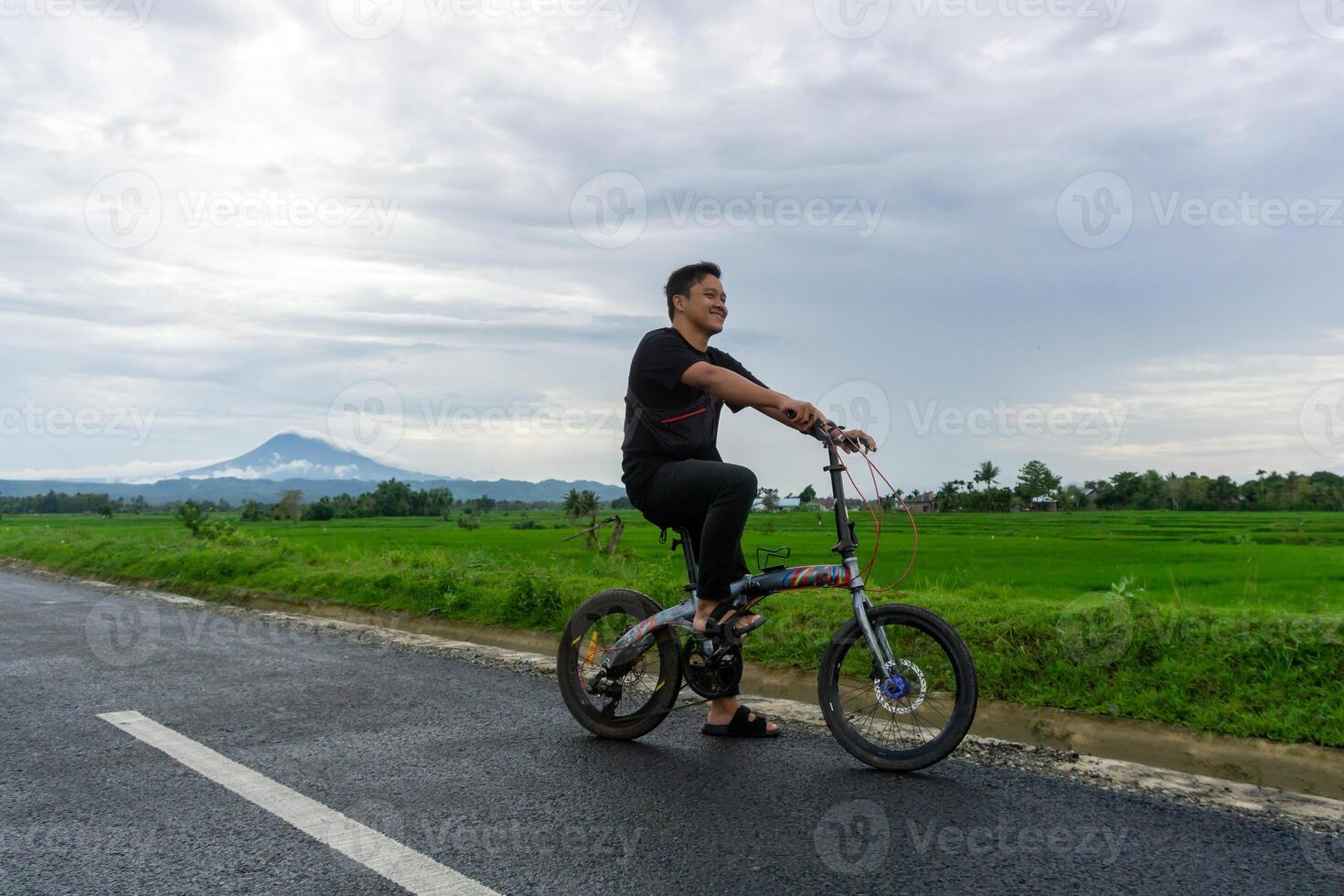 glücklich asiatisch Mann Reiten ein Fahrrad beim das Morgen auf das Asphalt Straße. Radfahren mit Berg und Paddy Reis Feld Aussicht beim das Hintergrund. foto