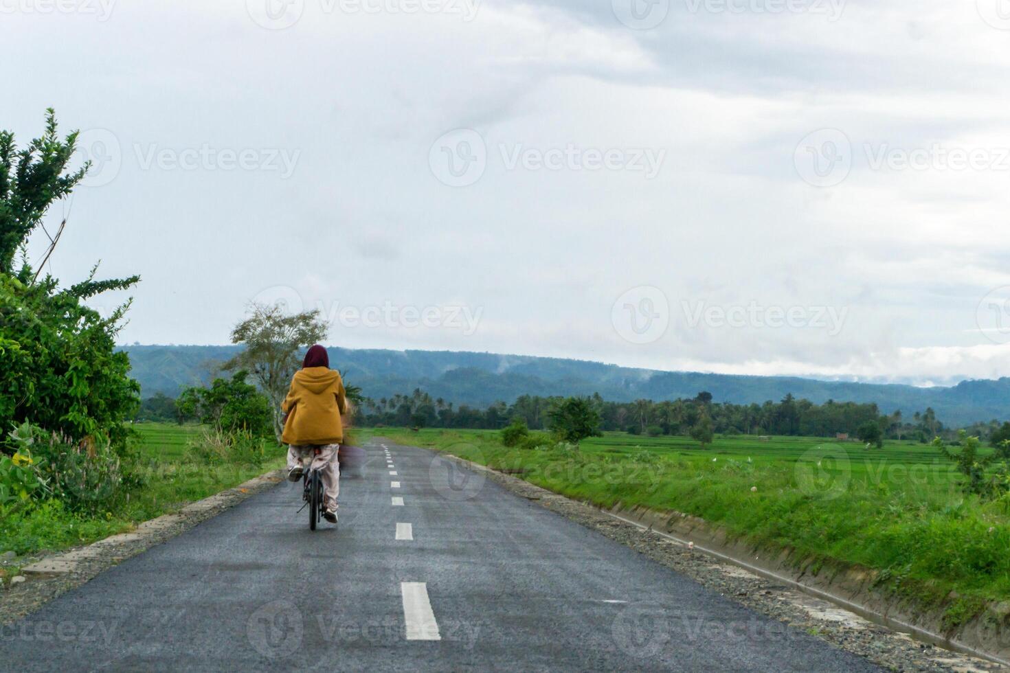 asiatisch Hijab Frau Reiten Fahrrad beim das Morgen auf das Asphalt Straße. ein Mädchen Radfahren mit Berg und Paddy Reis Feld Aussicht beim das Hintergrund. Kopieren Raum foto