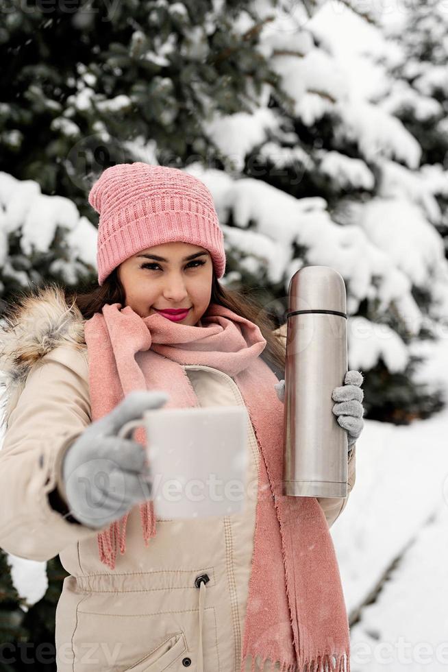 Schöne Frau in warmer Winterkleidung, die Thermoskanne hält und an einem verschneiten Tag im Freien heißen Tee oder Kaffee trinkt foto