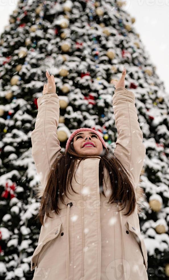 Frau in warmer Winterkleidung, die draußen am großen Weihnachtsbaum steht, Arme hoch, Schnee fällt foto