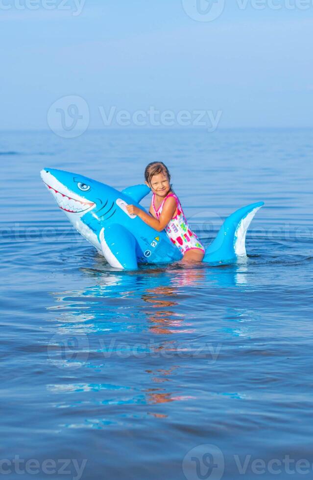 glücklich Mädchen von europäisch Aussehen Alter von 7 Schwimmen auf ein aufblasbar groß Hai Spielzeug im das Meer.Familie Sommer- Berufung Konzept. Vertikale Foto. foto