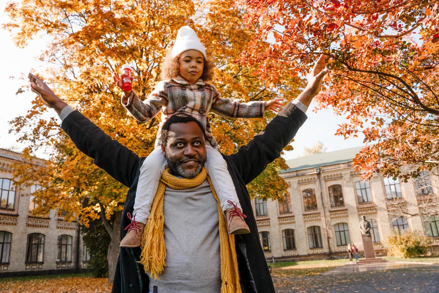 schwarzes Mädchen, das Spaß hat und im Herbstpark am Hals seines Großvaters sitzt foto