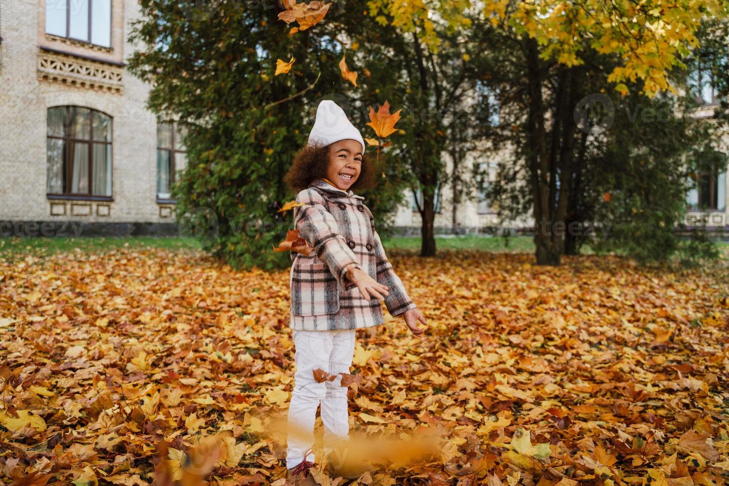 schwarzes Mädchen mit Mantel, das sich mit gefallenen Blättern im Herbstpark lustig macht foto