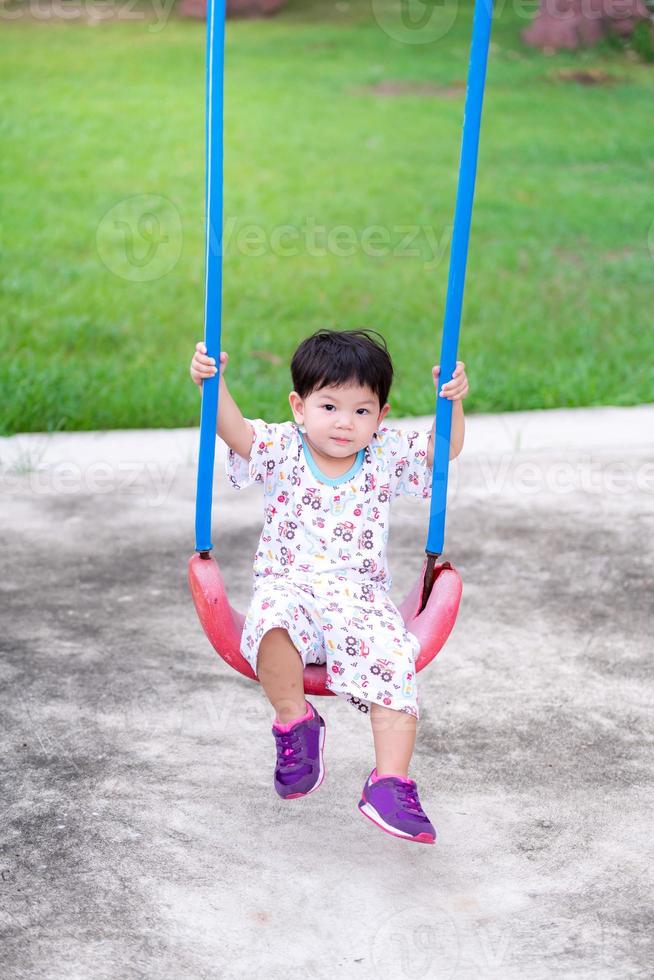 asiatischer Junge saß auf einer Schaukel und lächelte süß in die Kamera. Kinder spielen auf dem Spielplatz. Rund um den Spielplatz gibt es eine saftig grüne Wiese. Sohn ist 2 Jahre alt. foto